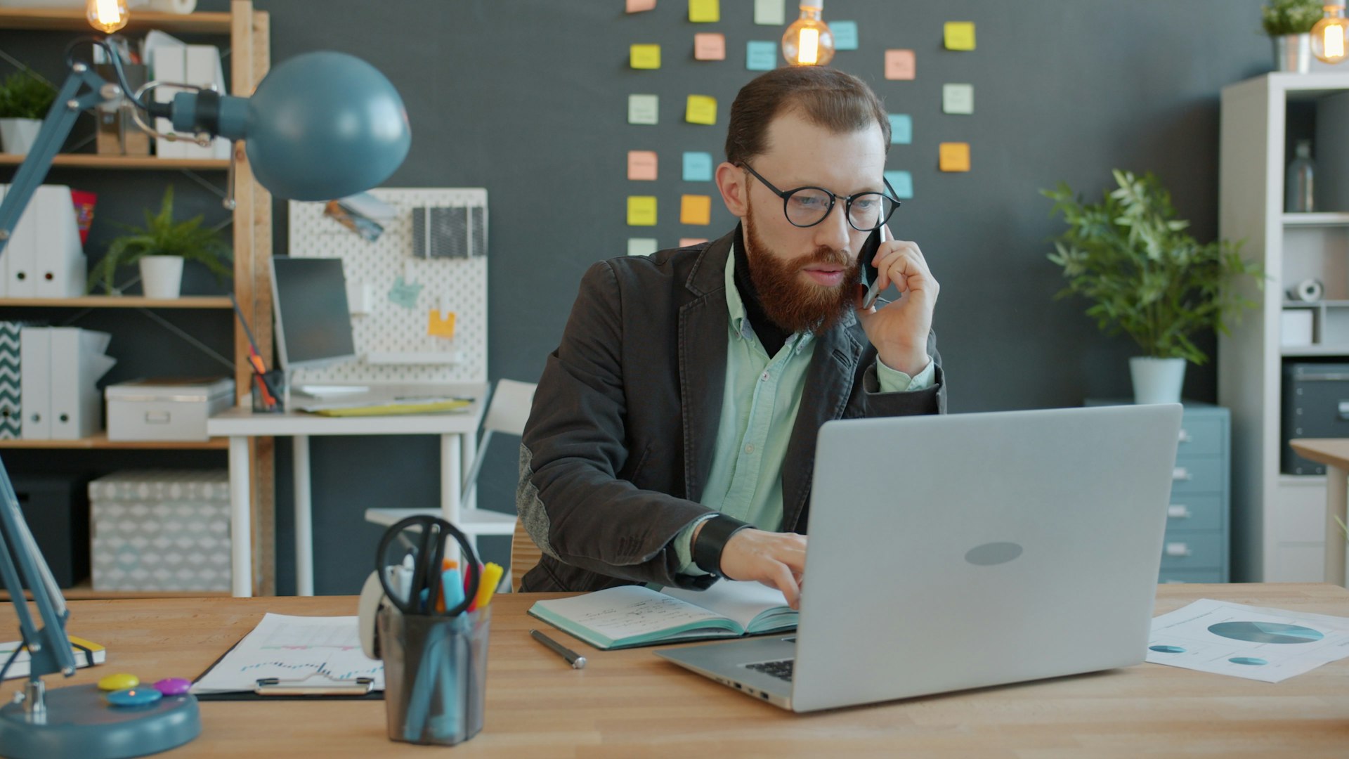 Man on phone working on laptop at desk.
